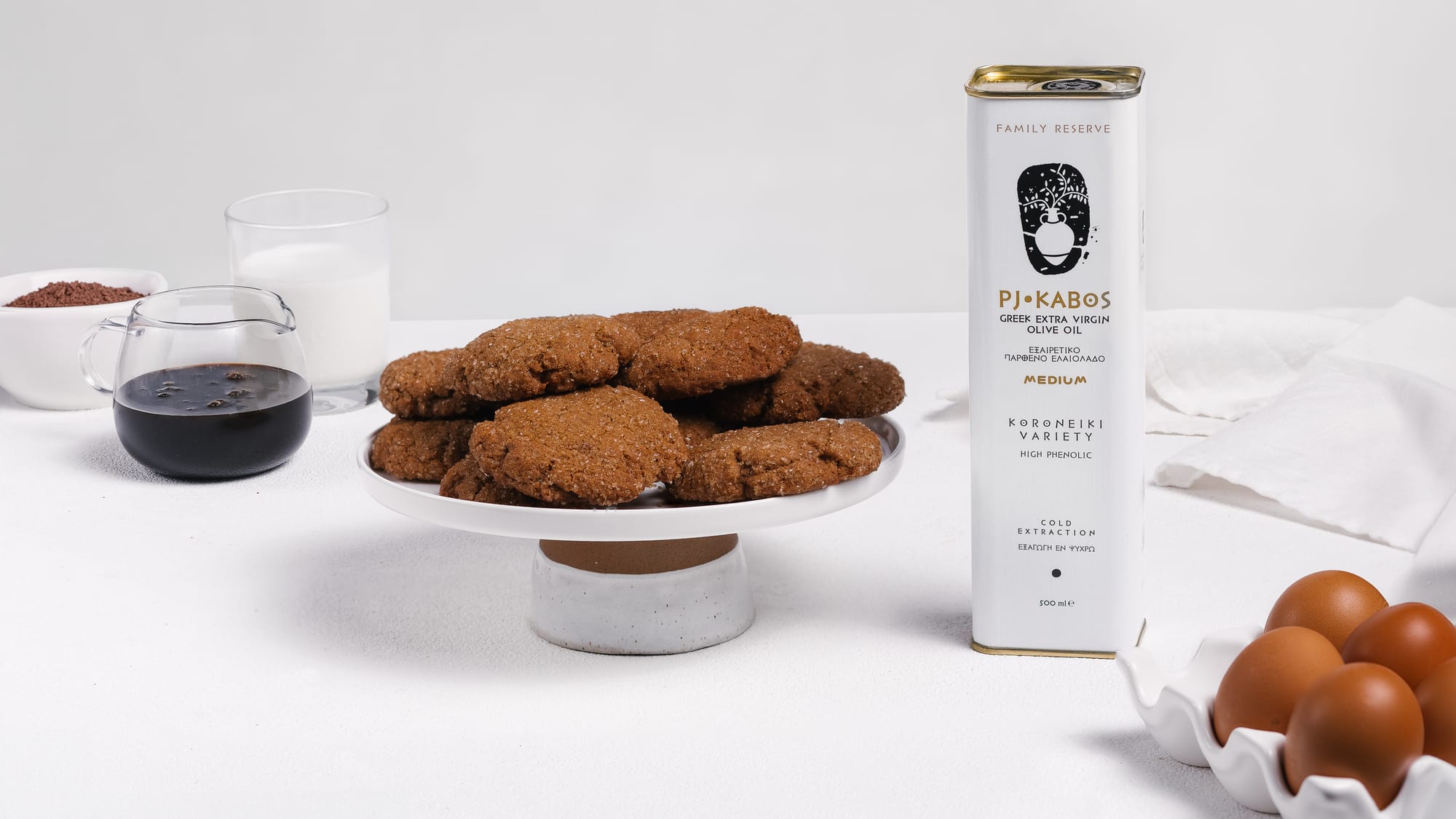 PJ KABOS Extra Virgin Olive Oil, eggs, flour, molasses, sitting on a tale around a plate of freshly baked ginger snap cookies.