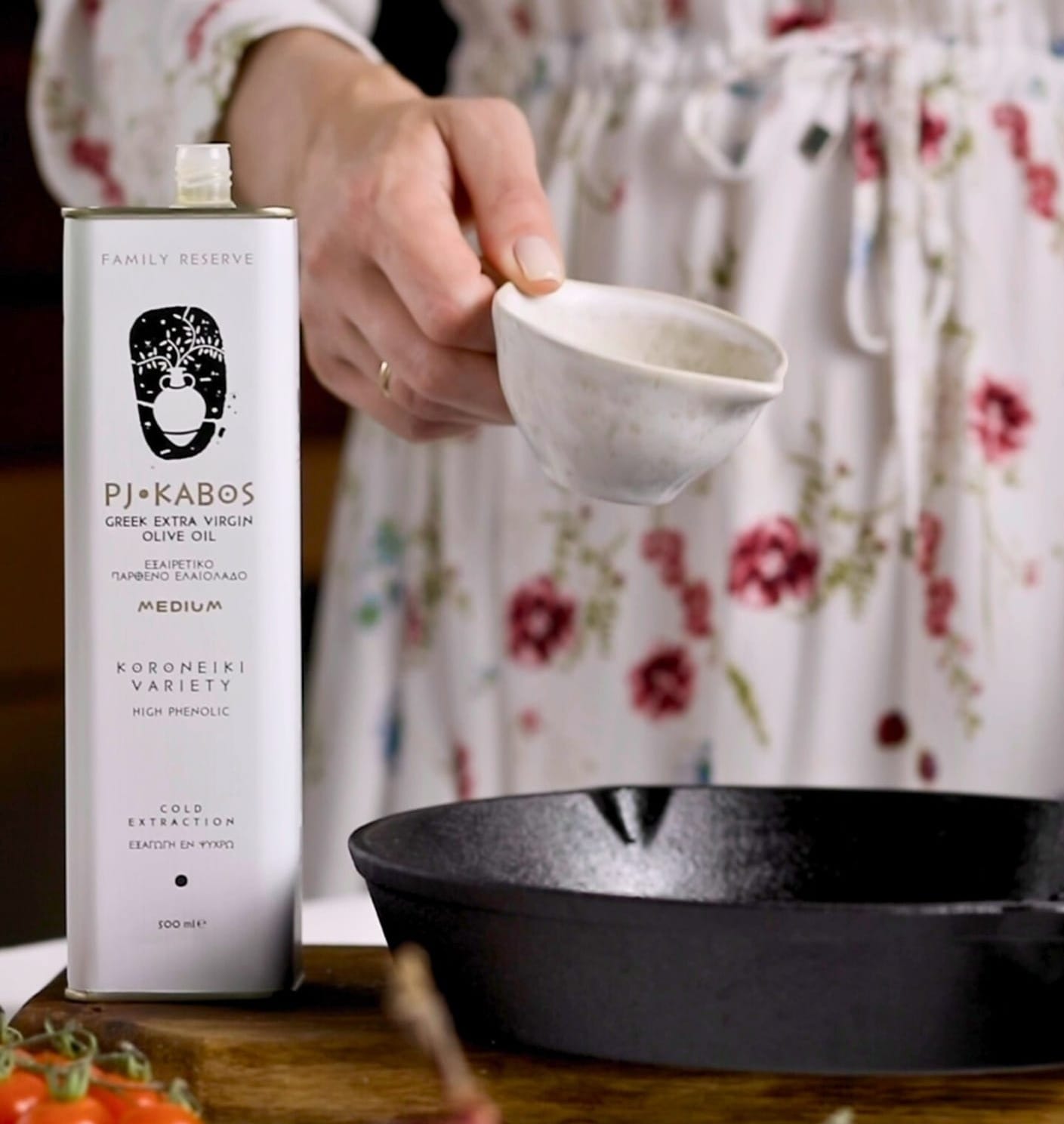 A lady is pouring olive oil into a pan. A tin of PJ Kabos Extra Virgin Olive Oil is on the table next to the cast iron skillet.