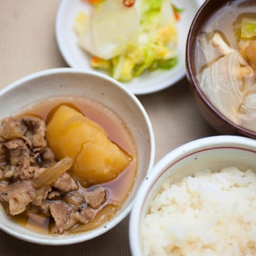 The Japanese dish of meat, potatoes and onion stewed in sweetened soy sauce called Nikujaga is shown along with rice and vegetables in additional bowls.