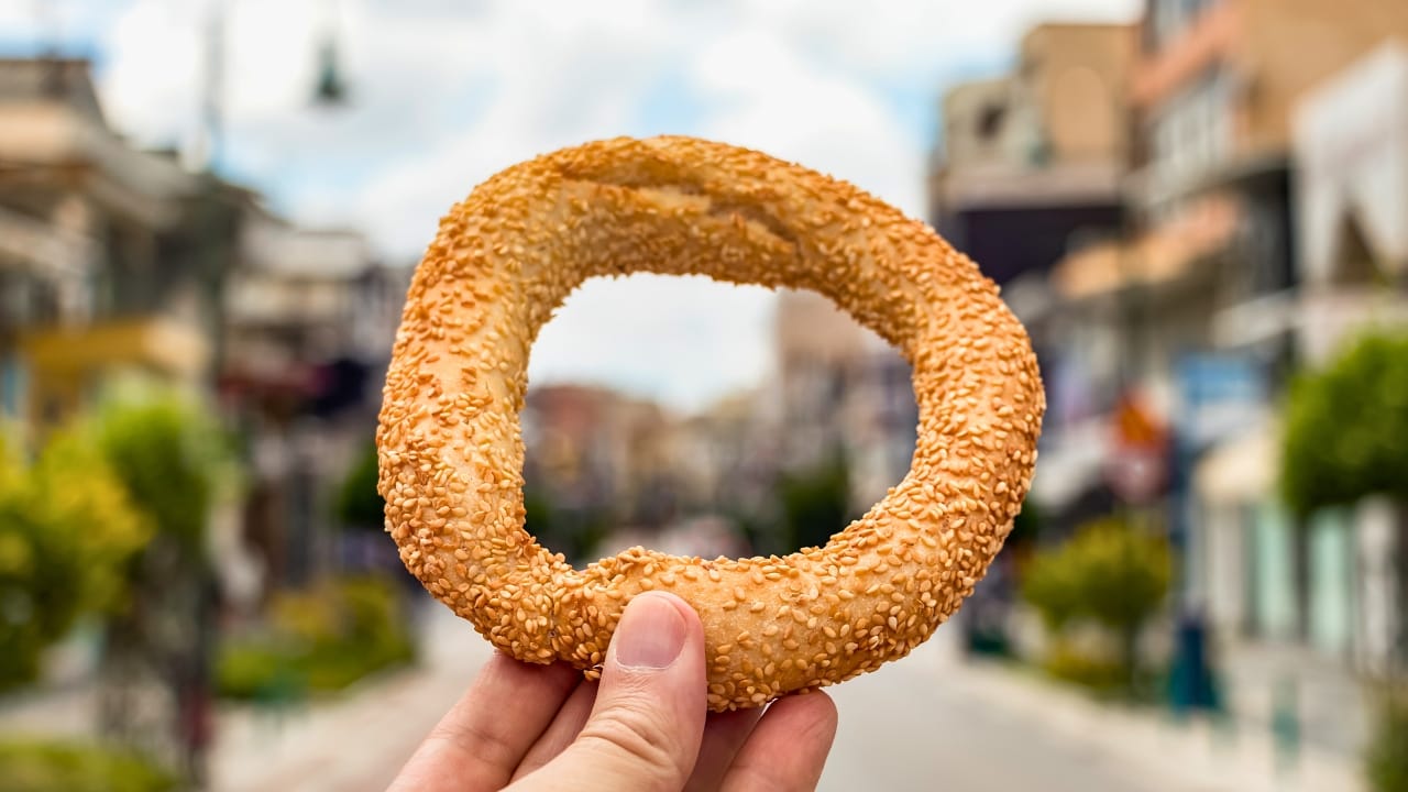 A person holding a Koulouri - an ancient ring roll covered in sesame seeds - above a modern Greek street.