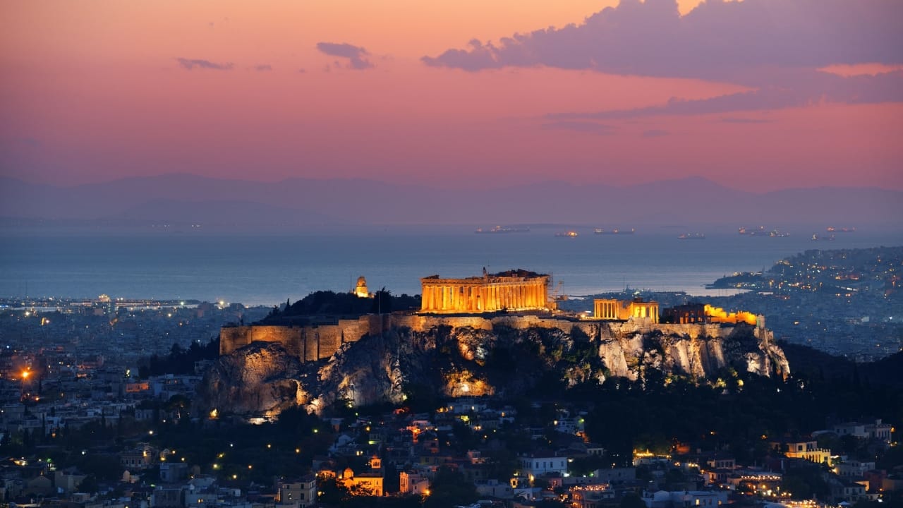 The Acropolis of Athens lit up at dusk with the city and sea below and around it.