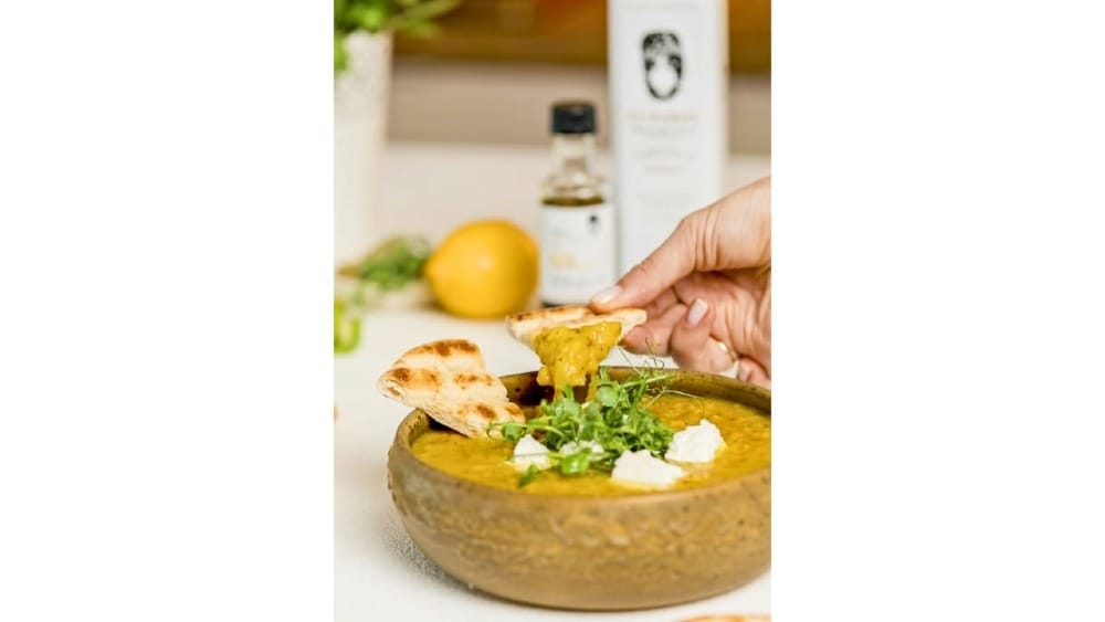 A bowl of PJ Kabos Yellow Split Pea Soup made with high-phenolic extra virgin olive oil, pita bread and the ingredients used to prepare it sits on a counter ready to be eaten.