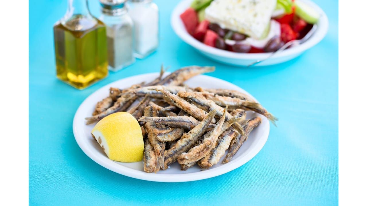 Gavros Tiganitos (Fried Anchovies)sitting on a table with a Greek salad and olive oil